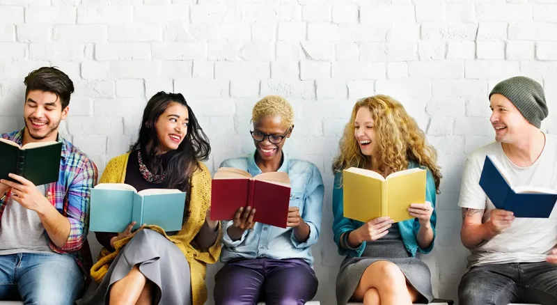 Five people sitting against a white brick wall, laughing as they read colourful books